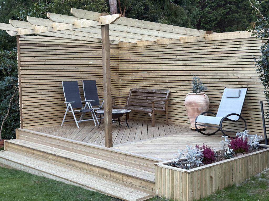 A timber decking, with a pergola and a slatted fence on two sides. The decking has a built in flower bed on one edge and the other has steps leading down into the garden.