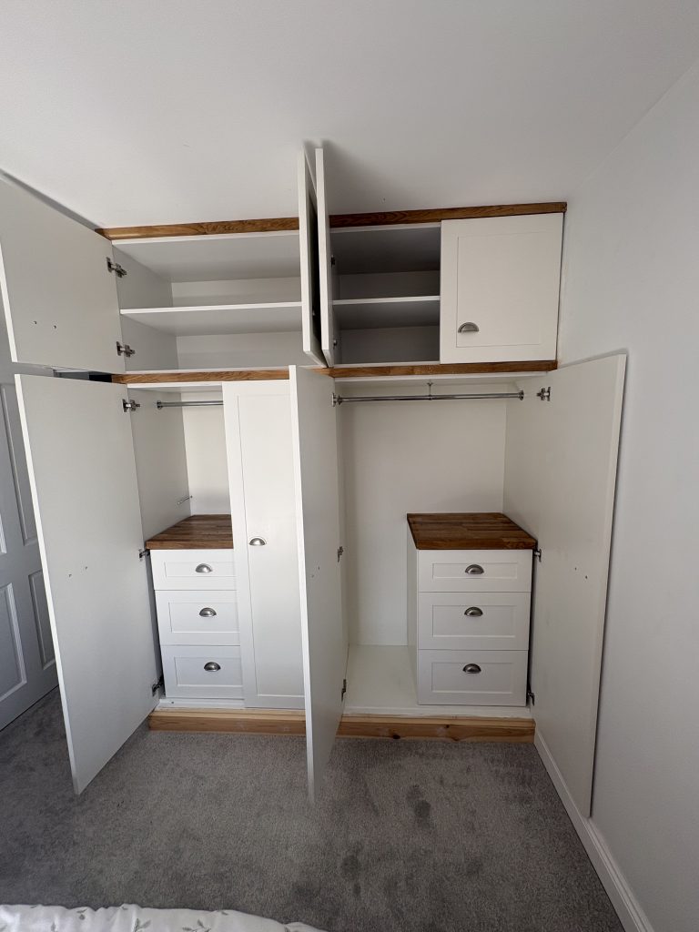 A white built in wardrobe with doors open. Showing the storage space at the top and the cupboard space below. Below there is a unit with three drawers inside the cupboard. The drawers have a timber work top.